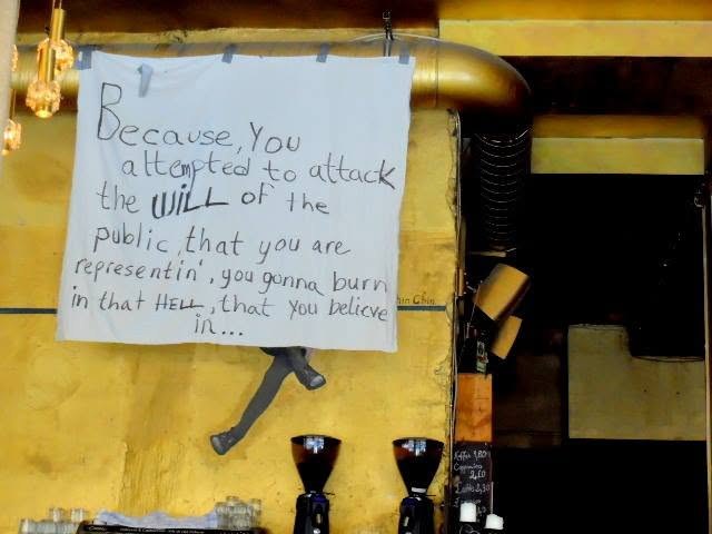 hand-lettered on a bedsheet above the coffee grinders &mdash; because you attempted to attack the will of the public that you are representin&rsquo;, you gonna burn in that hell that you belive in. spelling left in. a caf&eacute; that hung a curse where the menu goes.
