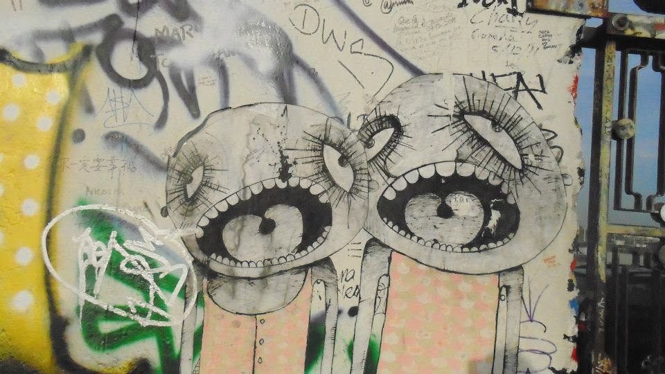 two heads, all eye and all teeth, drawn in pen on the wall between other people s signatures. the eyes are enormous because in a surveilled city looking is the loudest thing you can do.