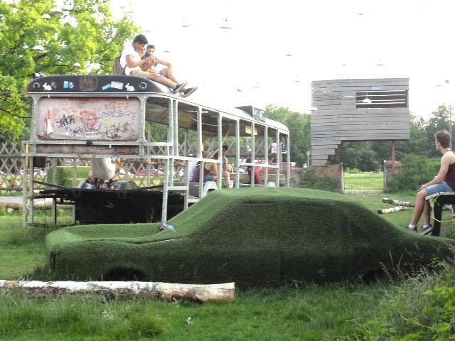 a car buried under turf, a stripped bus turned into a grandstand, on land nobody had bought yet. the machine goes under the grass; the people sit on the wreck. this is what free land looks like before it gets a price.
