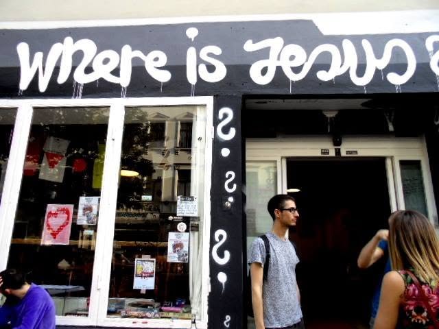 painted across the shopfront backwards, so it reads from the inside: where is jesus? three question marks under it, in case you thought it was rhetorical. two people wait at the door, not looking up.