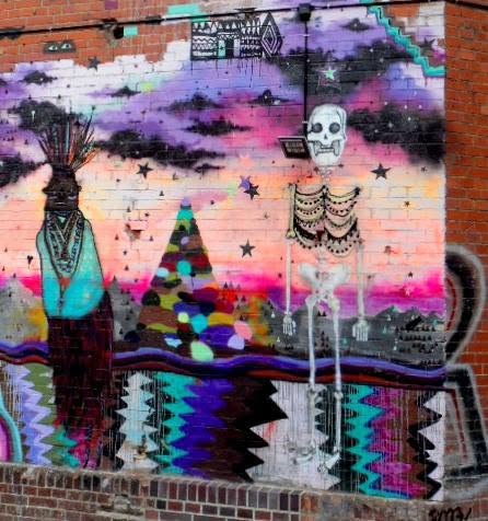 a skeleton in a sunset, hung with beads, facing a figure whose whole head is feathers. day-of-the-dead grammar on a berlin firewall: the dead dressed up and standing in the light instead of tidied away.