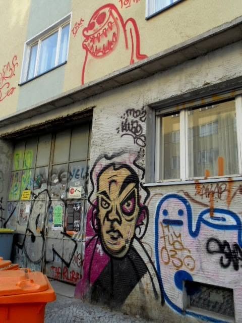 a red thing screaming at roof level, a furious man in a pink collar at street level, someone&rsquo;s blue blob between them. four artists, one wall, no agreement. the bins are the only thing anyone maintains.