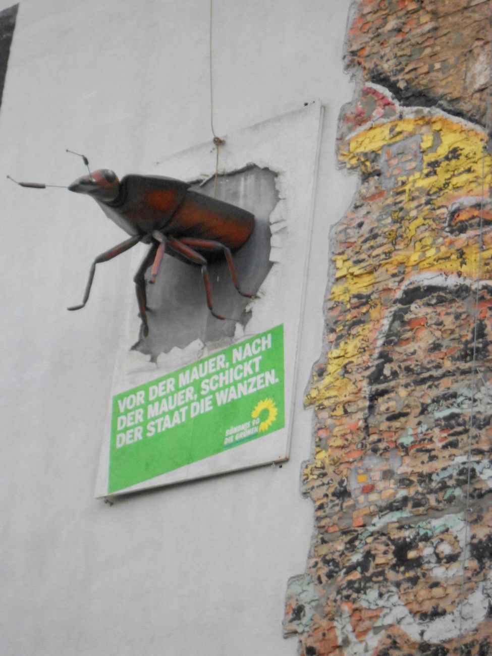 a beetle the size of a dog bursting out of the plaster, and under it a green-party poster: vor der mauer, nach der mauer, schickt der staat die wanzen. before the wall, after the wall, the state sends the bugs   wanzen means both the insect and the wiretap. two germanies, one habit.