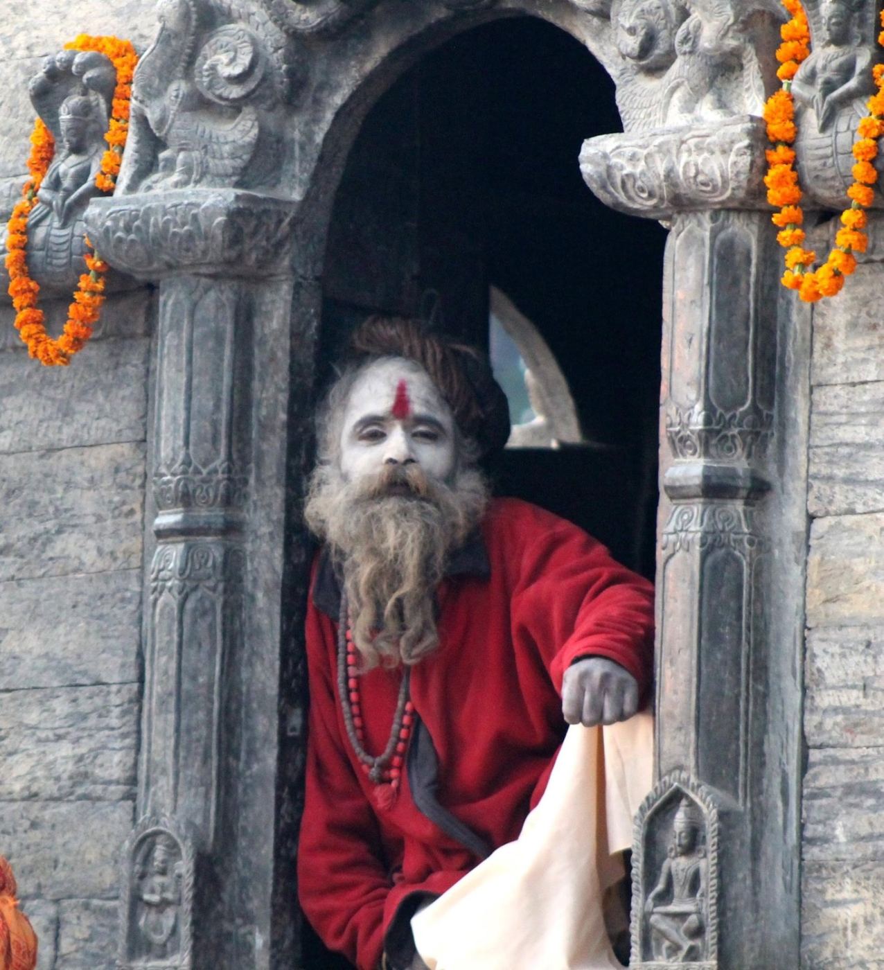 a sadhu in a stone doorway hung with marigolds, red robe, white ash