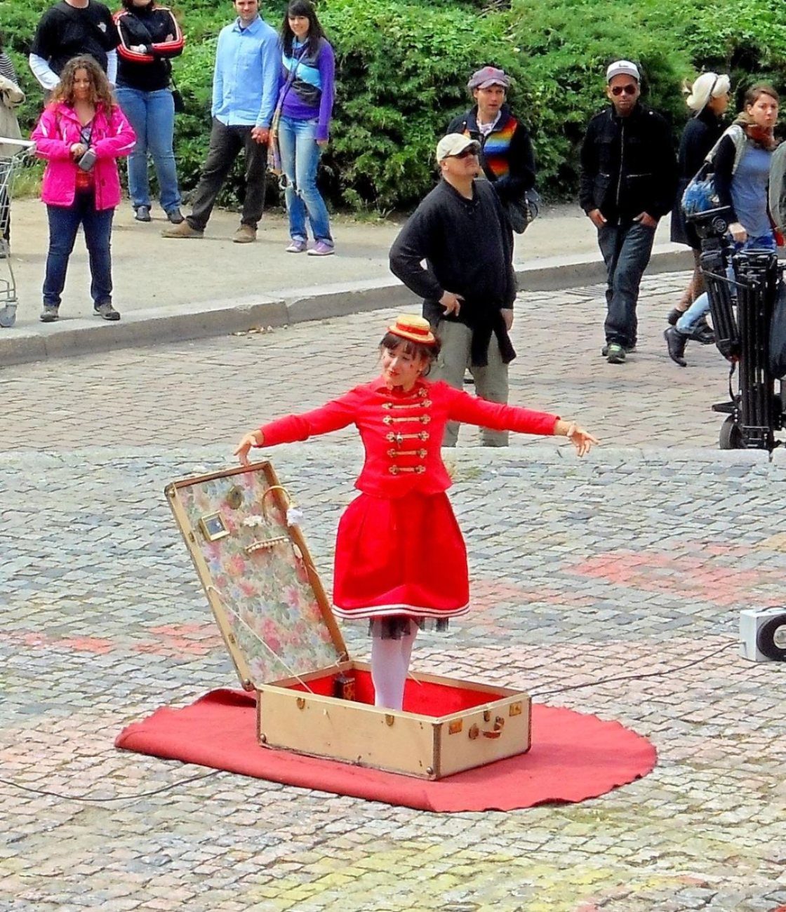 a puppet in a red dress dances in her own suitcase while the crowd walks past.