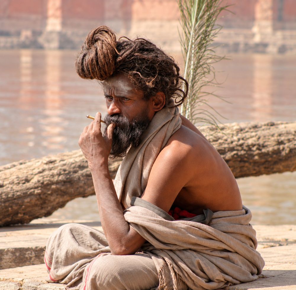 A sadhu with a high knot of matted hair smoking beside the river