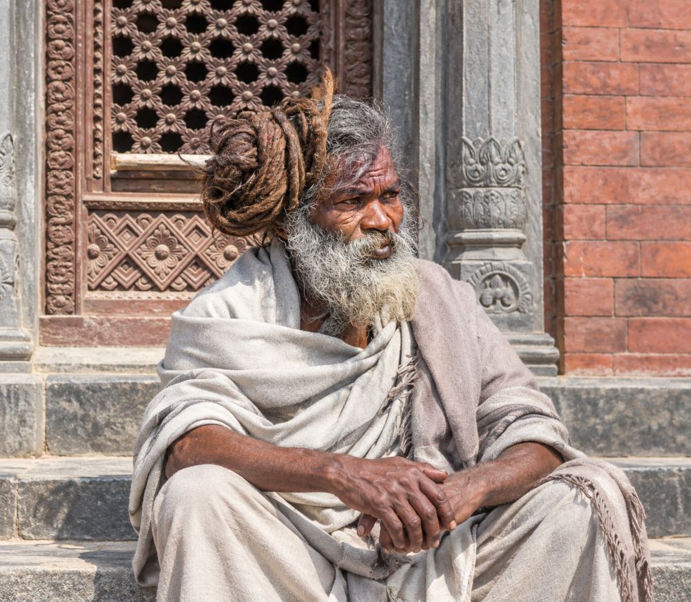An old sadhu with grey matted hair coiled high, wrapped in a pale shawl