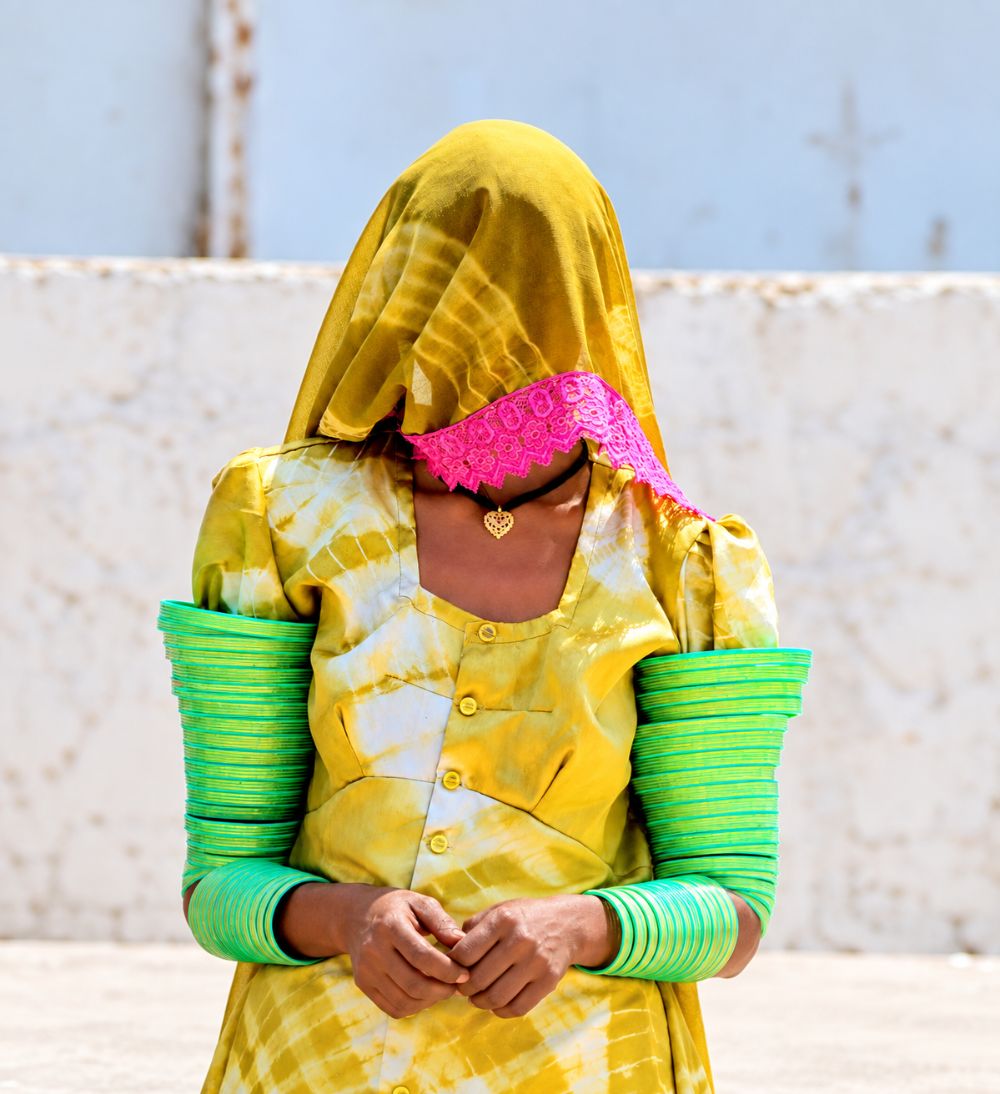 A woman fully veiled in yellow tie-dye, both arms stacked with green bangles