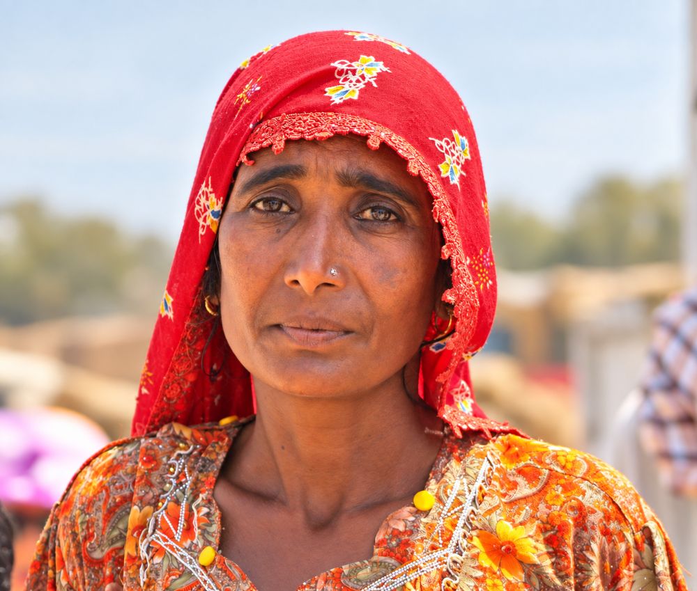 An older woman in a red printed dupatta edged with lace, village blurred behind