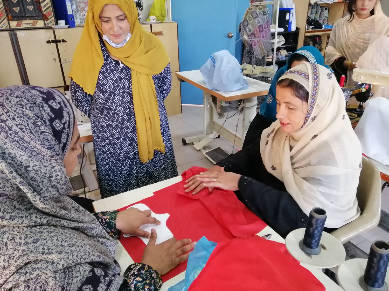 Meera guides a seated trainee cutting red fabric